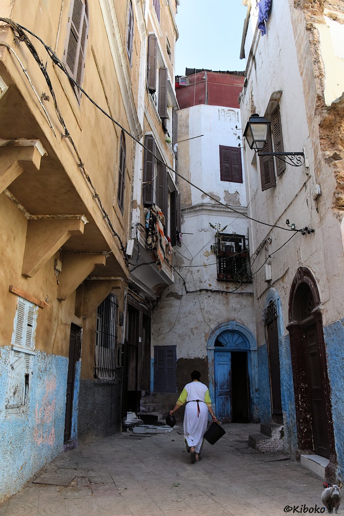 Das Bild zeigt eine Hochformataufnahme, die nach einem Haus nach rechts abbiegt. Die Gebäude haben 3-4 Geschosse. Eine Frau mit weißem Kittel läuft in die Gasse.