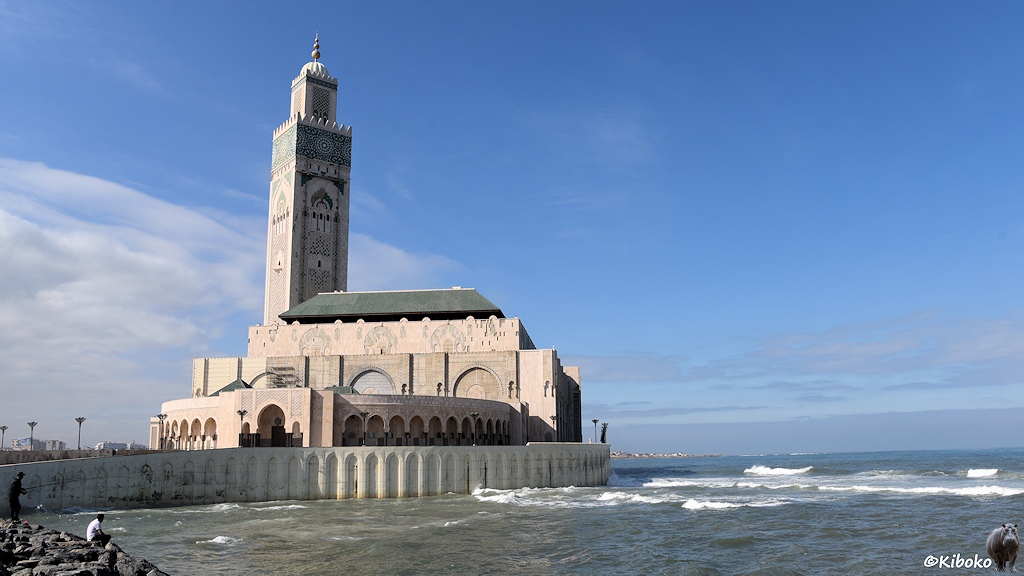 Das Bild zeigt eine Moschee aus hellem Steinen mit einen hohen, viereckigen Turm. Eine Schutzmauer it ornamenten schützt die Moschee vor den Wellen des grünlichen Meeres. Im Vordergrund sind zwei Angler auf einer Felsbarriere am Meer.
