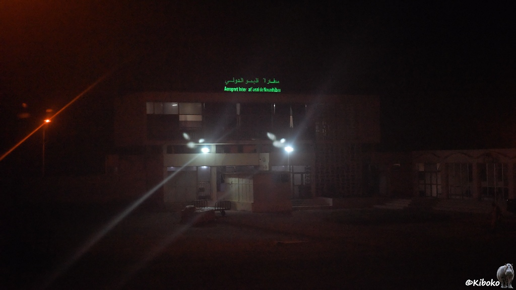 Das Bild zeigt eine Nachtaufnahme eines kleinen zweistöckigen Flughafengebäudes aus Beton. Auf dem Gebäude ist eine grüne Leuchtschrift: Aeroport Inter at onal de Nouadhibou. Vor dem Terminal stehen zwei Laternen. Dahinter leuchtet eine Straßenlaterne mit rötlichem Licht. Vor dem Gebäude stehen zwei Gepäckkarren.
