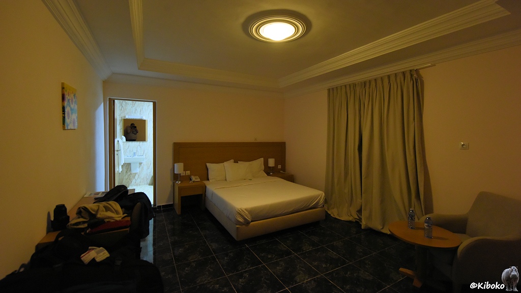 Das Bild zeigt ein Hotelzimmer mit Doppelbett, schwarzen Fußbodenfliesen und Stuckdecke.