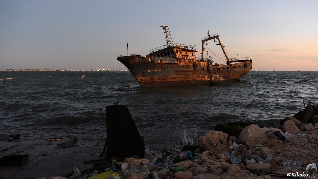 Das Bild zeigt einen verrosteten Fischtrawler in der Morgendämmerung. An der Horizontlinie sind beleuchtete Gebäude. Im Vordergrund mit felsigem Strand, der mit Plastikflaschen vermüllt ist.