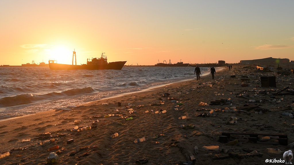 Das Bild zeigt zwei Menschen, die im Gegenlicht der Morgensonne am Strand entlanglaufen. Der Strand ist liegen viele weiße, grüne und gelbe Plastikflaschen, die von der tiefstehenden Sonne von hinten angestrahlt werden und dadurch leuchten.