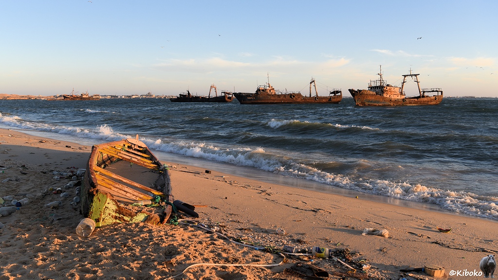 Das Bild zeigt ein Beiboot aus Holz von schräg hinten. Das Boot liegt am Strand zwischen Plastikflaschen. Im Hintergrund dümpeln verrostete Fischtrawler.