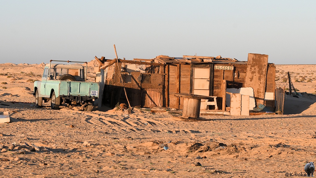 Das Bild zeigt ein Strandhaus aus Holzplanken. Davor steht ein Tisch aus einer leeren Kabelrolle. Links neben dem Haus steht ein alter, hellblauer Land Rover. Drumherum ist Wüste.