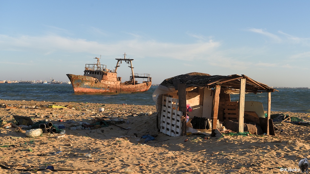 Das Bild zeigt eine einfache Hütte aus angeschwemmten Baumaterialien am Strand. Im Hintergrund dümpelt ein verrosteter Fischtrawler in der Bucht.