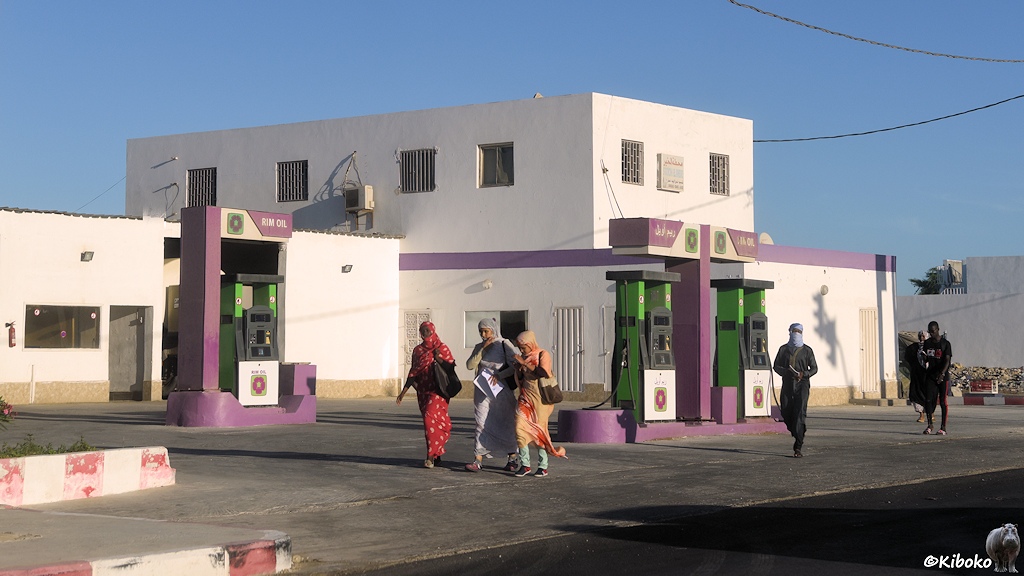Das Bild zeigt eine Tankstelle ohne Dach mit drei Zapfsäulen. Darüber steht Rim Oil. Die Sockel und Pfeiler sind violett angemalt. Im Hintergrund ist ein zweistöckiges Gebäude mit Flachdach und violetten Zierstreifen. Vor der Tankstelle laufen drei verschleierte Frauen in bunten Kleider und zwei Männer.