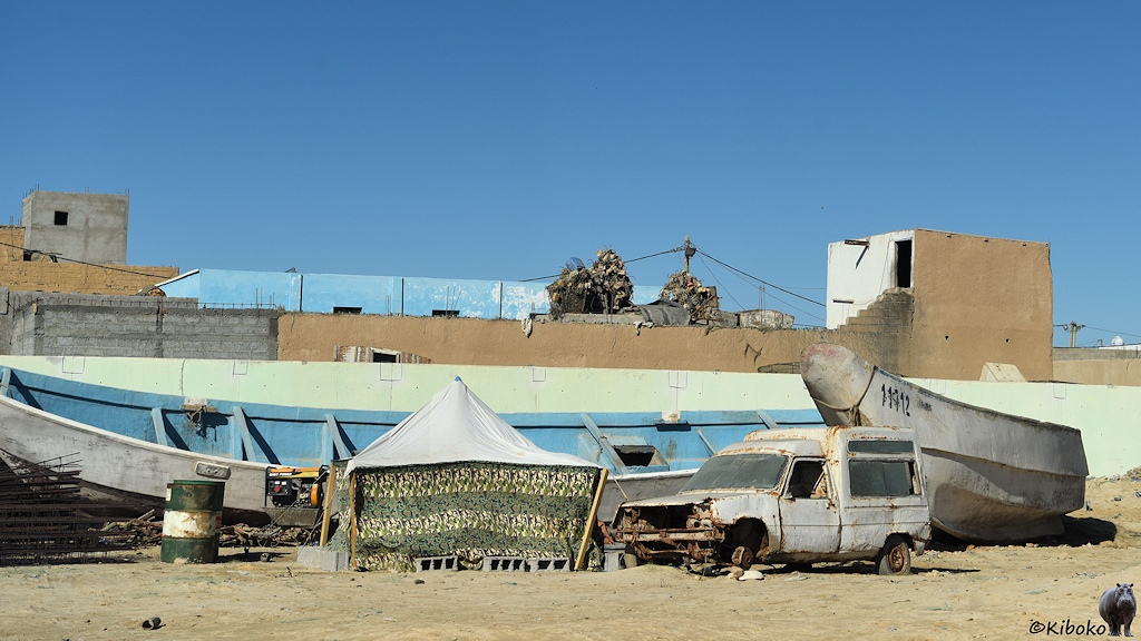 Das Bild zeigt ein viereckiges, grünes Zelt mit weißem Dach. Rechts daneben steht ein ausgeschlachteter Lieferwagen ohne Räder. Dahinter liegen zwei offene Boote auf dem Sand. Im Hintergrund ist eine hellbraune Mauer über die die Dächer von Häusern schauen.