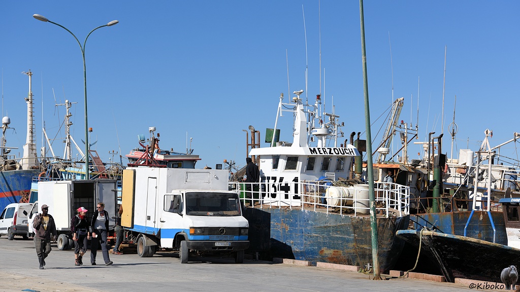 Das Bild zeigt den Kai in einem Fischereihafen. Am Kai stehen zwei Kleinlaster mit Kastenaufbau und geöffneten Ladetüren. Am Kai liegt ein Fischerboot mit verkratzten dunkelblauen Rumpf und weißen Aufbauten. Dahinter liegen weitere Fischerboote im Hafen.