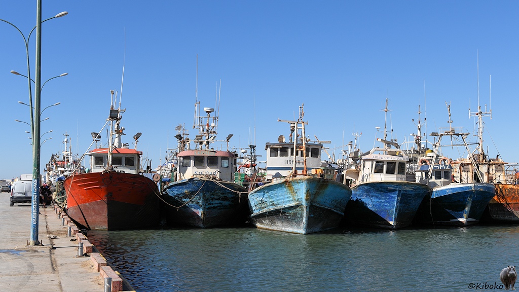 Das Bild zeigt die Bugspitzen von einer Reihe Fischerboote. Das Erste liegt links am Kai und hat einen roten Rumpf. Die folgenden vier Schiffe haben einen Rumpf in unterschiedlichen Blautönen.