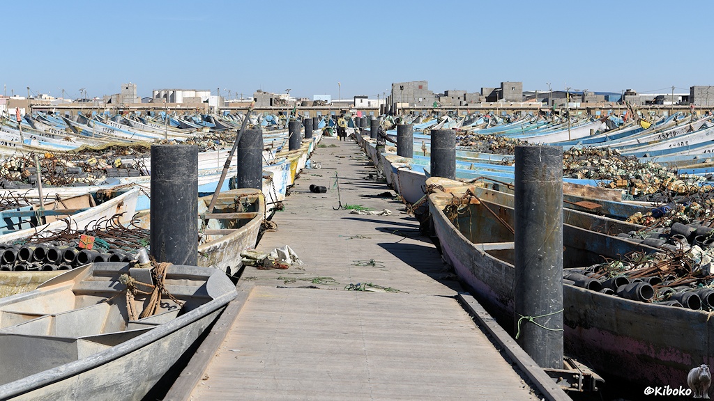 Das Bild zeigt einen Pier aus Holz im Hafen. Auf beiden Seiten sind kleine graue, weiße und hellblaue Fischerboote festgemacht. In den Fischerbooten liegen schwarze Schwimmer und verrostete Metallhaken.