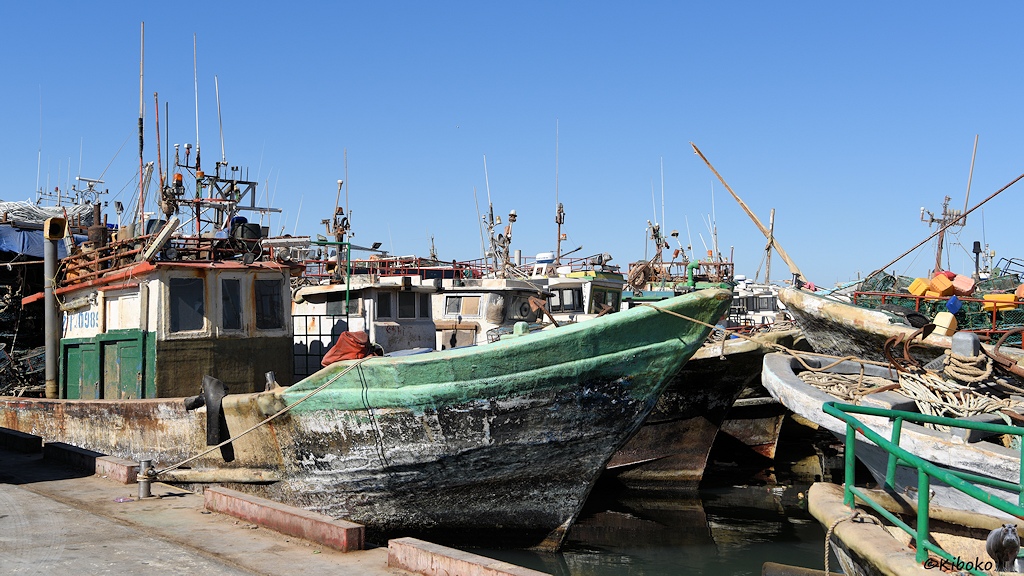 Das Bild zeigt eine Reihe von Fischerbooten mit Steuerstand. Am Kai liegt ein Boot mit schwarzem Rumpf und hellgrüner Reeling. Dahinter schauen die Bugsitzen und Hecks weitere Fischerboote.