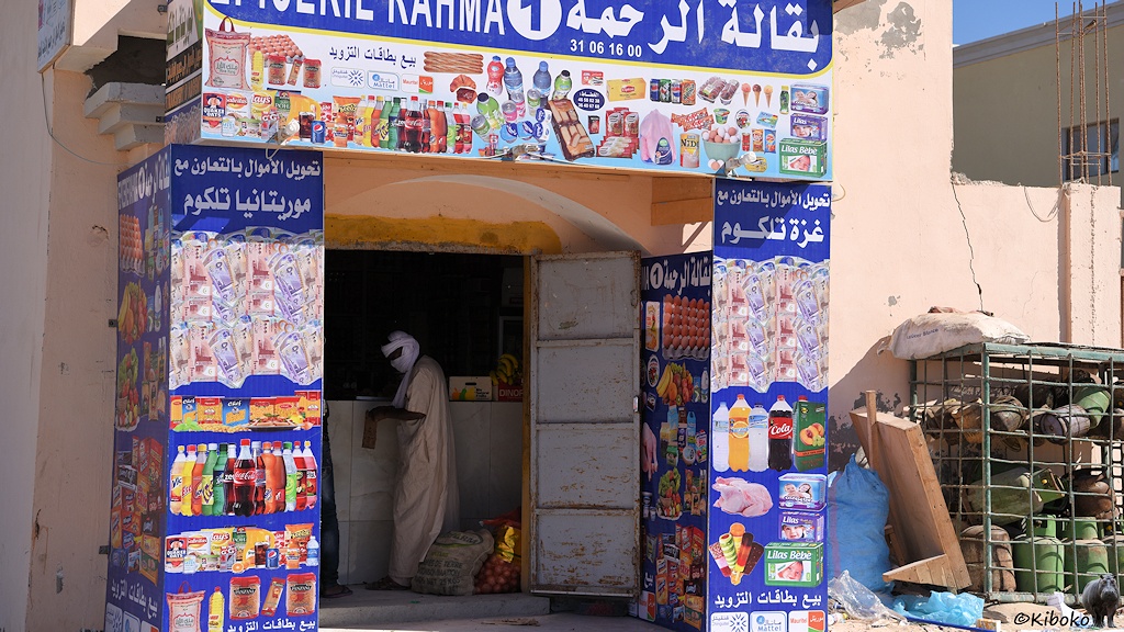 Das Bild zeigt ein kleines Geschäft mit einer offenen Stahltür. Darin ist ein Kunde mit weißen Turban und beigem Gewand. Rund um die Tür ist ein blaues Schild mit Cola Flaschen, Keksen, Geldscheinen, Eis und Babywindeln.