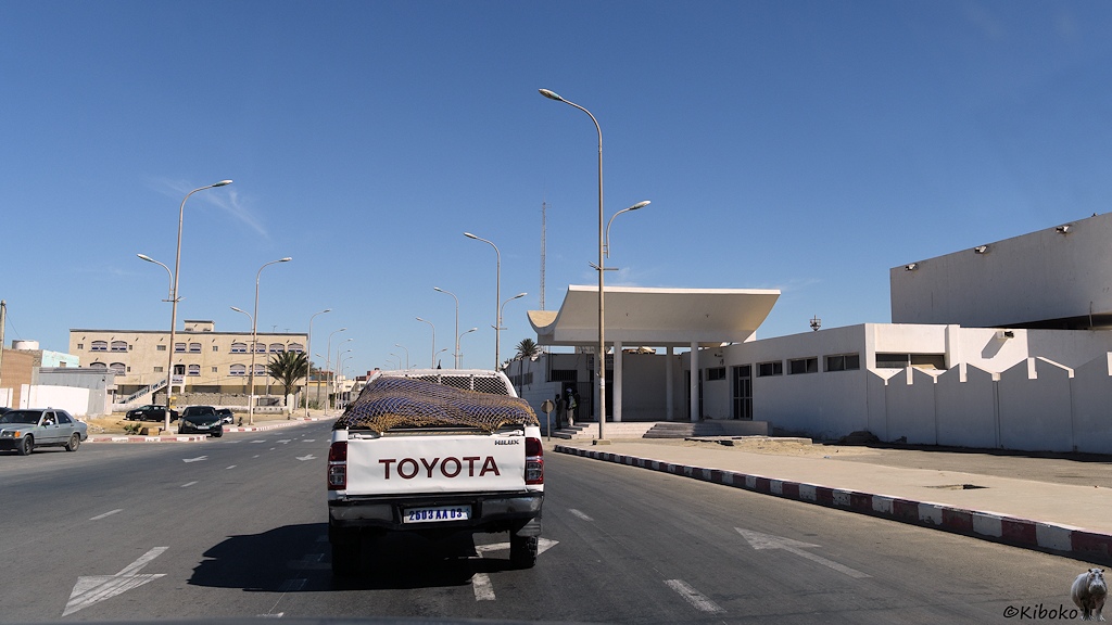 Das Bild zeigt das Heck eines vorausfahrenden Toyota Pickup auf einer vierspurigen Straße. Am rechten Bildrand ist ein einstöckiges weißes Gebäude mit einen geschwungenen Dach auf vier Säulen.