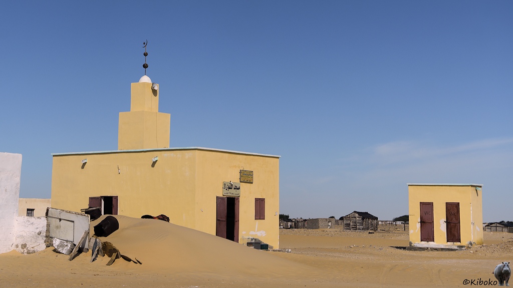 Das Bild zeigt eine kleine, viereckige, gelbe Moschee mit braunen Holztüren und Fensterläden. Sie hat einen kleinen viereckigen Turm. Daneben steht ein kleines gelbes Klohäuschen mit zwei Türen. Vor der Moschee ist ein hoher Sandhaufen. Im Hintergrund sind einfache Holzhütten.