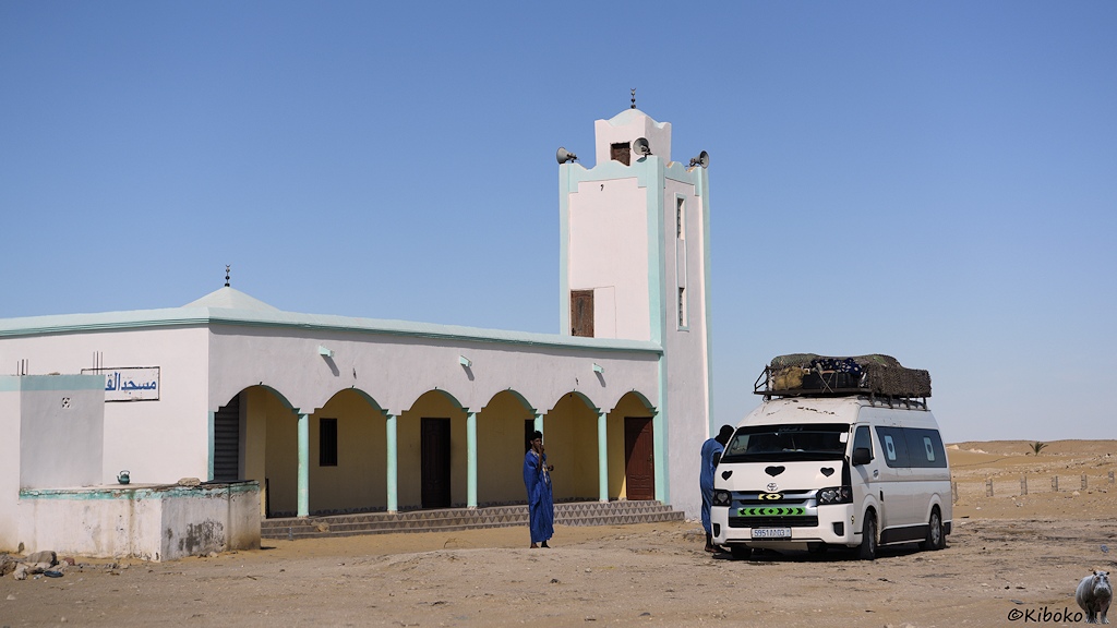 Das Bild zeigt eine weiße Moschee mit Säulengang im Eingangsberich. Sie hat ein viereckiges Minarett mit hellblauen Ecken. Vor der Moschee stehen zwei Männer im blauen Gewandt und ein weißer Minibus mit Dachladung.