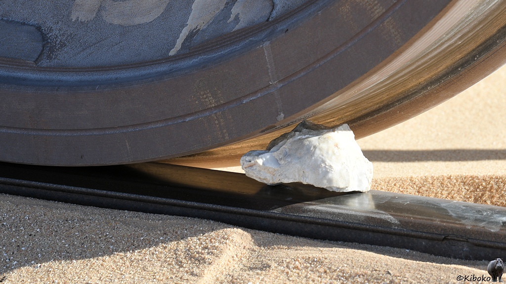 Das Bild zeigt einen keilförmigen, weißen Stein, der zwischen Schiene und Eisenbahnrad eingeklemmt ist.