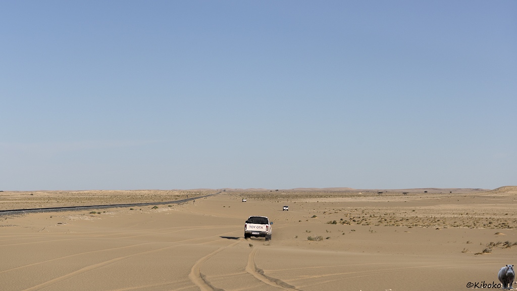 Das Bild zeigt einen Geländewagen von hinter der durch die Wüste fährt. Links dabeben verläuft die Bahnstrecke. Im Hintergrund sind ganz klein zwei weitere Geländewagen.