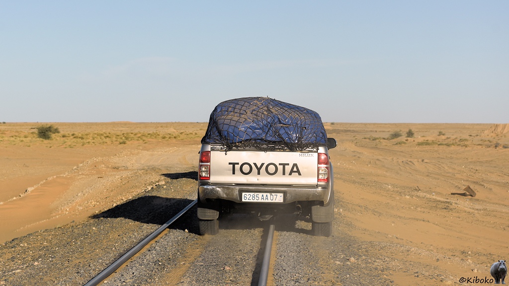 Das Bild zeigt einen vorausfahrenden Geländewagen von hinten, der auf den grauen Schotter der Bahngleise fährt. Die linken Räder fahren zwischen den Gleisen. Die rechten Räder rollen rechts von den Gleisen. Voraus ist eine weite Ebene in der Wüste.