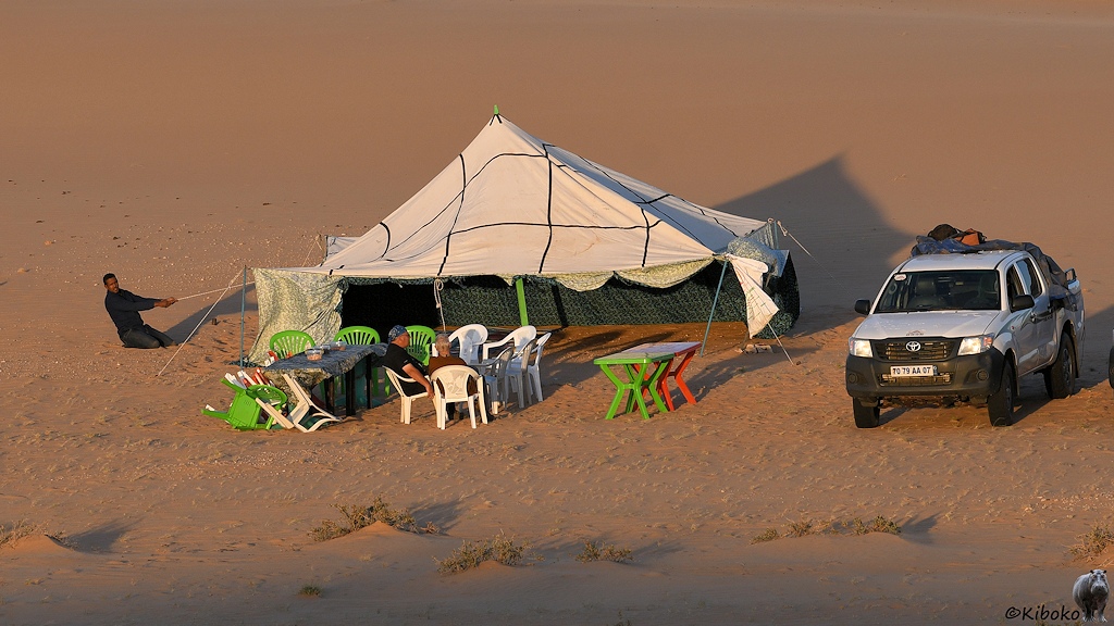 Das Bild zeigt ein großes viereckiges Zelt, an dem ein Mann gerade ein Seil spannt. Vor dem Zelt sind weiße und grüne Plastikstühle. Rechts daneben steht ein Geländewagen.