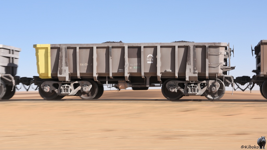 Das Bild zeigt einen vierachsigen, offenen, graubraunen Güterwagen mit einem gelben Ende und massiven vertikalen Streben. Das Bild ist ein Mitzieher. Die Wüstenlandschaft ist verwischt.