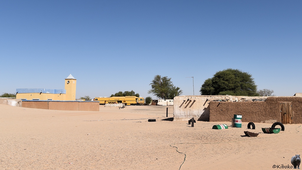Das Bild zeigt die Ortsmitte. Links ist eine gelb angemalte Moschee mit Blechdach und viereckigen Turm, die mit einer braunen Lehmmauer umgeben ist. Rechts ist ein Hauseingang mit einer braunen Mauer. Davor liegen grün-weiße Fässer und Autoreifen im Sand. Eine Stromleitung liegt auf dem Boden und führt zum Haus. Im Hintergrund steht eine gelbe Maschine am Bahnhof.