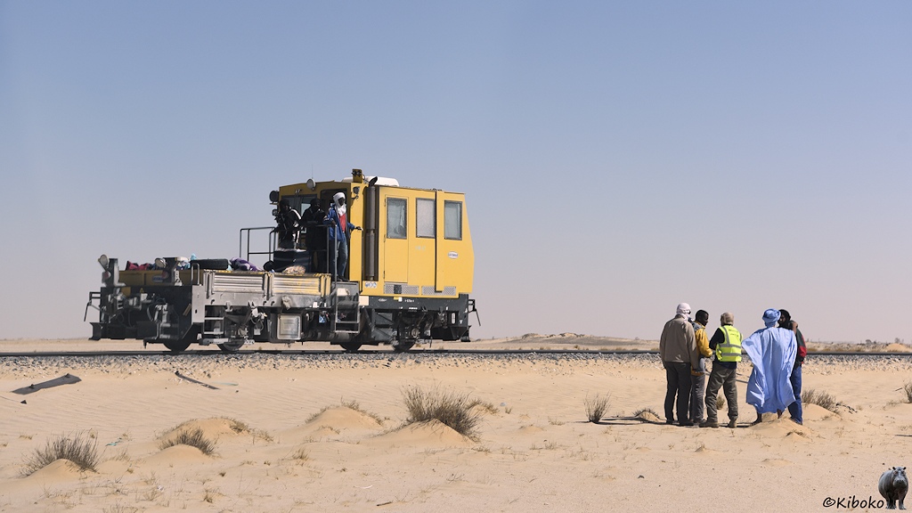 Das Bild zeigt eine zweiachsige Draisine mit einer kantigen, gelben Führerkabine und einer beladenen Plattform. Vor der Draisine stehen fünf Männer im Wüstensand.