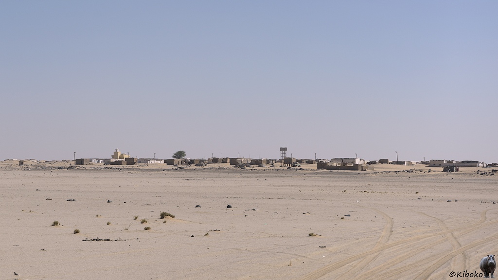 Das Bild zeigt eine Panoramaufnahme von einem Ort in der Wüste. Die Gebäude sind einstöckige Steinbauten mit Flachdach. Eine Moschee mit gelben Turm und ein aufgeständerter Wassertank überragen die Häuser.