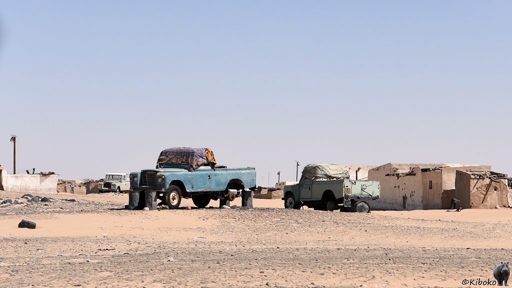 Das Bild zeigt einen dunkelblauen und einen blassgrünen Land Rover Pickup auf einem unbefestigten Platz zwischen einstöckigen Steinhäusern. Die Autos stehen auf schwarzen Stahlfässern. Bei beiden Autos fehlt das linke Hinterrad. Die Fahrerkabinen sind durch Decken geschützt.