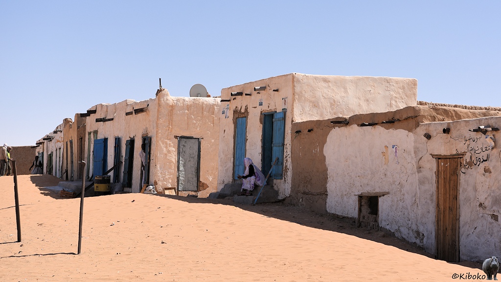Das Bild zeigt eine Reihe kleiner wüfelförmiger, weißer Lehmhäuser mit blauen Türen. Vor den geöffneten Türen sitzen Verkäufer im Schatten.
