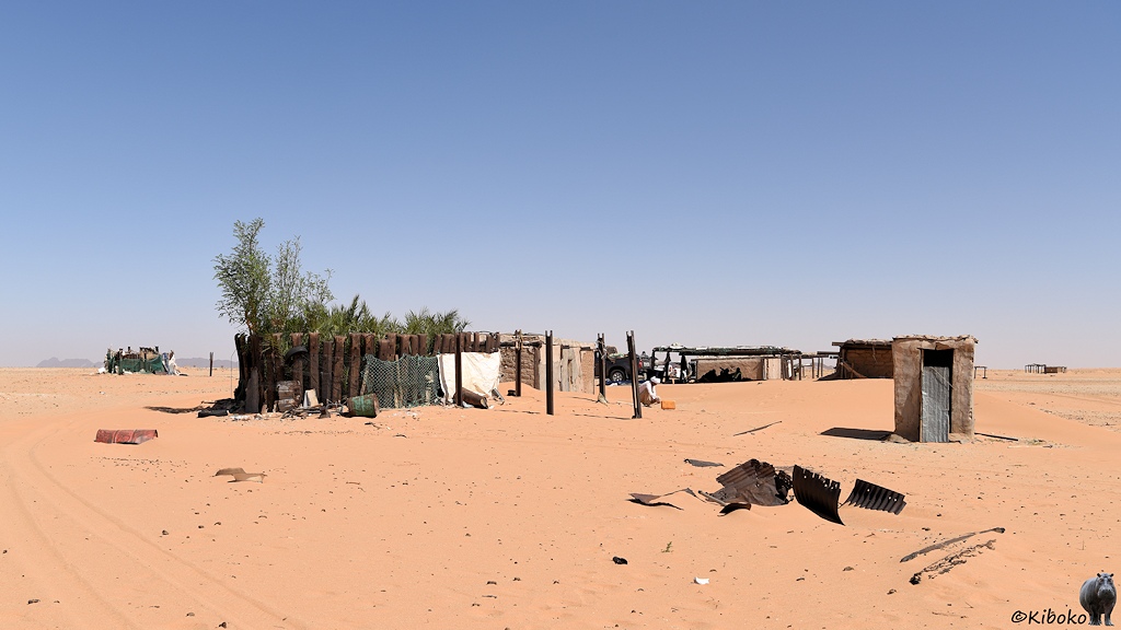 Das Bild zeigt ein paar Hütten in der Wüste. Link ist ein kleiner Garten mit einem Zaun aus senkrecht stehenden Stahlschwellen. Darüber ragen die Äste von Bäumen. In der Bildmitte hockt ein Mann, der einen gelben Kanister füllt.