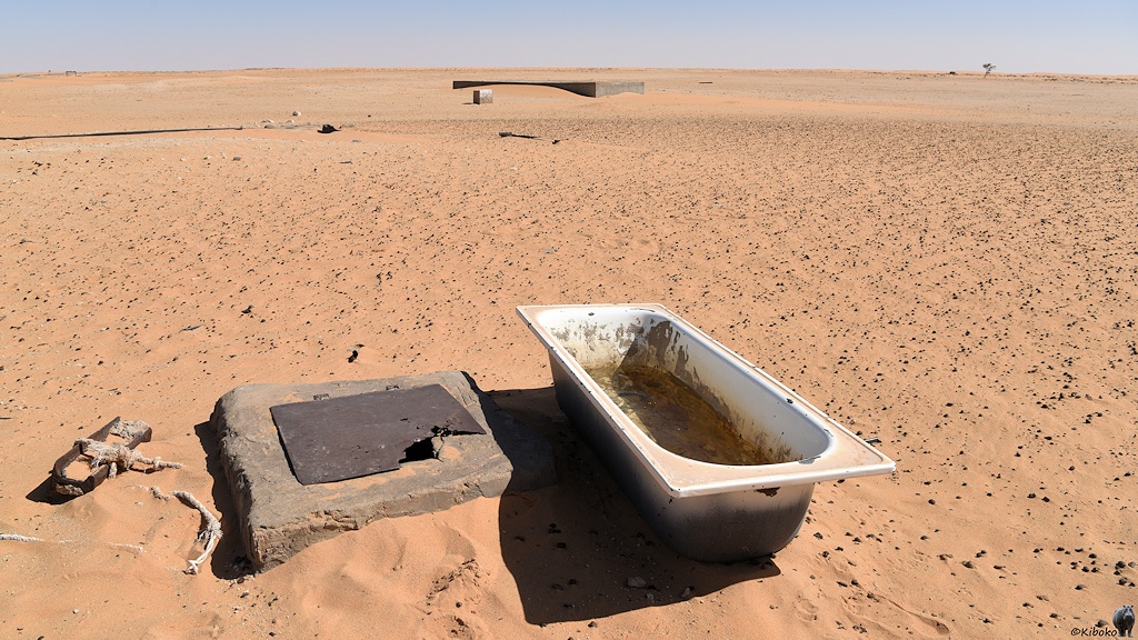 Das Bild zeigt eine leere Badewanne in der Wüste. Links daneben ist ein Betonfundament mit einer Öffnung. Die ist durch eine rostige Klappe abgedeckt. Rund um die Badewanne sind kleine dunkle Kugeln im blass lachsfarbenen Sand.