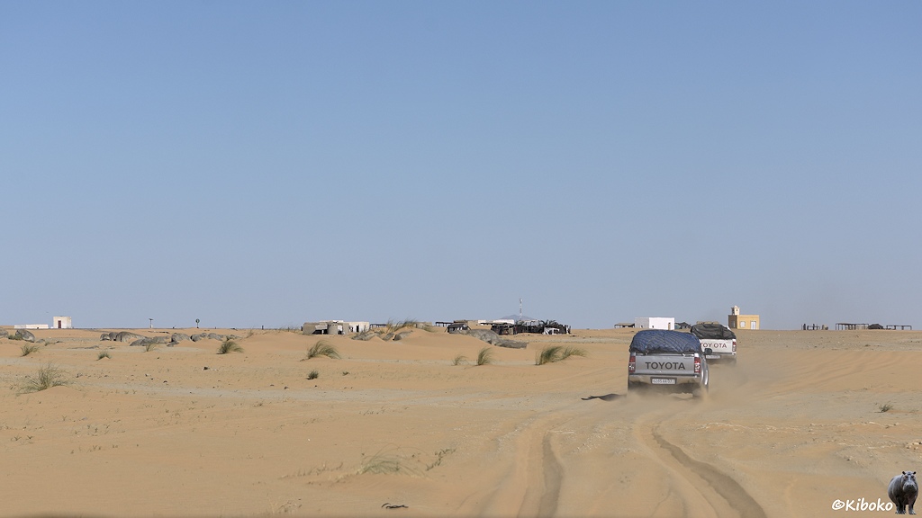 Das Bild zeigt mehrere Geländewagen fahren hintereinander auf eine kleine Ortschaft in der Wüste zu. Die Autos hinterlassen eine tiefe Spur im Sand.
