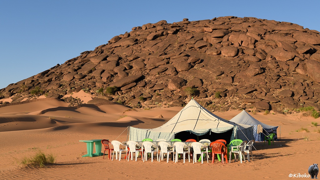 Das Bild zeigt zwei rechteckige Zelte vor einem Hügel aus rotbraunen Felsen. Davor steht eine Tafel aus Plastiktischen mit weißen, roten und grünen Plastikstühlen im Wüstensand.
