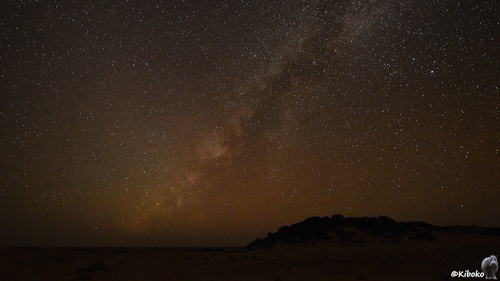 Das Bild zeigt die Milchstraße über der Wüste. Die Milchstraße verläuft schräg von links unten über die Bildmitte nach oben. Drum herum leuchten unzählige Sterne. Durch den Staub in der Luft schimmert der Himmel orange. Am Boden zeichnet sich rechts ein felsiger Hügel gegen den Sternenhimmel ab.