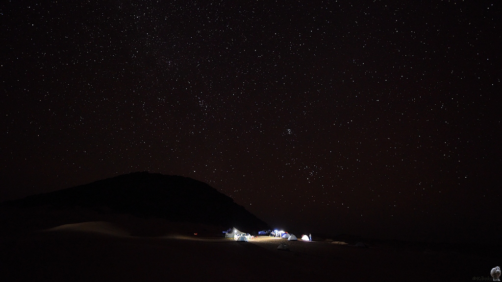 Das Bild zeigt beleuchtete Zelte unter einem Sternenhimmel. Ein Berg hinter dem Camp zeichnet sich schwach gegen die Sterne ab.