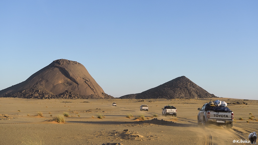 Das Bild zeigt vier Geländewagen bei der Fahrt durch eine beige Wüste. Die Autos nähern sich einen hohen Berg aus blankem rotbraunem Fels. Rechts daneben ist ein kleinerer, dunkler Felsen.