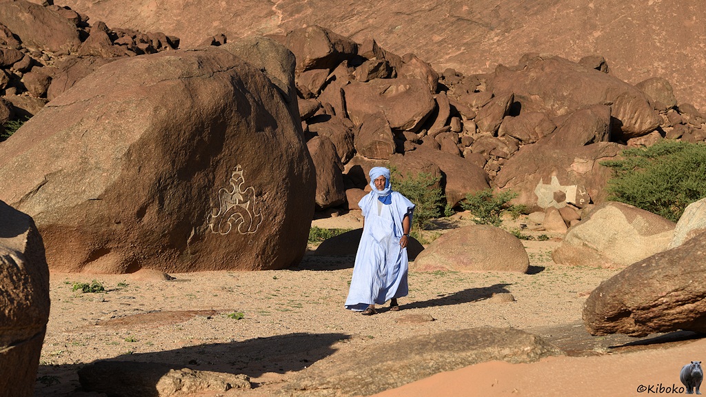 Das Bild zeigt einen Mann im hellblauen langen Gewandt beim Vorbeigehen an einen rötlichen Felsen mit einem eingravierten Kunstwerk. Im Hintergrund sind Haufen von kleineren Felsen und ein paar kleine Bäume.