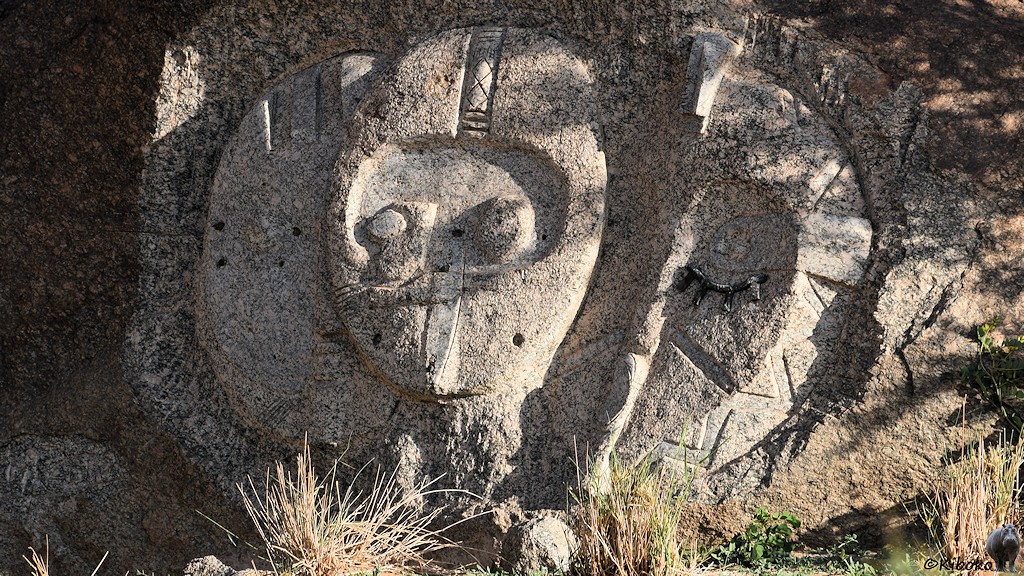 Das Bild zeigt eine ovale Bildhauerarbeit mit einem Gesicht im Zentrum an einen größeren Felsbrocken.