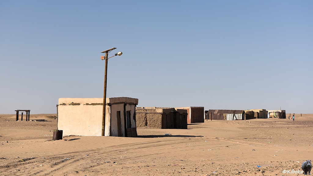 Das Bild zeigt eine Ansammlung von würfelförmigen Lehmhäusern in der Wüste. Vor dem ersten Haus steht ein Holzmast mit einer Laterne. Auf dem Mast sind Solarzellen und eine Batterie montiert.