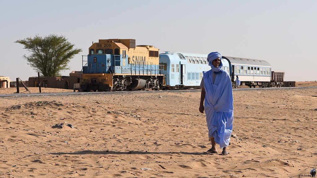 Das Bild zeigt einen älteren Mann in einem hellblauen Gewandt. Dahinter steht ein Zug mit einer beige-blauen Diesellok, einen hellblauen doppelstöckigen Wagen, eine blau-weißen Personenwagen und einen Erzwagen.