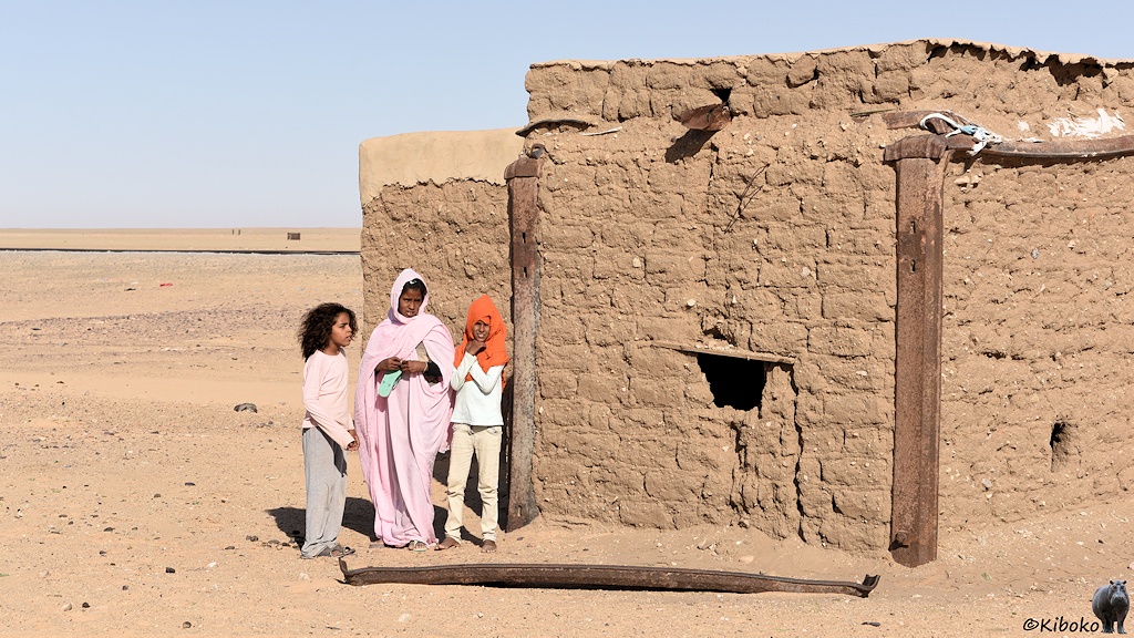 Das Bild zeigt ein Lehmhaus mit einem Guckloch als Fenster in der Wüste. Davor stehen eine Frau und zwei Mädchen. Die Frau ist in einem rosafarbenen Umhang gehüllt.
