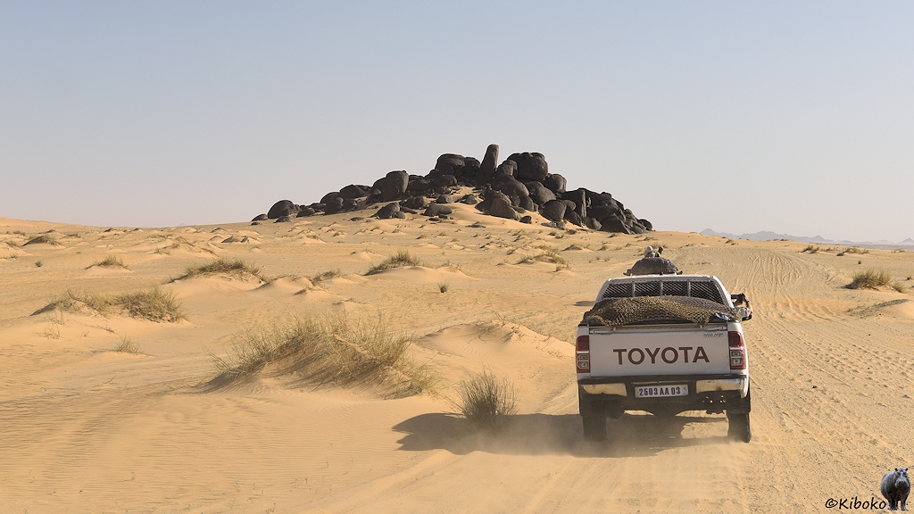 Das Bild zeigt einen vorausfahrenden Geländewagen in einen Dünenfeld aus gelblichen Sand. Auf den Dünenspitzen sind Grasbüschel. Am Horizont ist ein Hügel aus schwarzen Felsen.