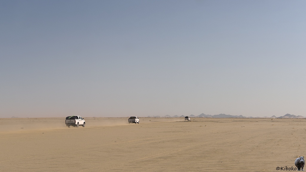 Das Bild zeigt drei Geländewagen, die auf einer ebenen Wüstenfläche auf eine Bergkette in der Ferne zusteuern.