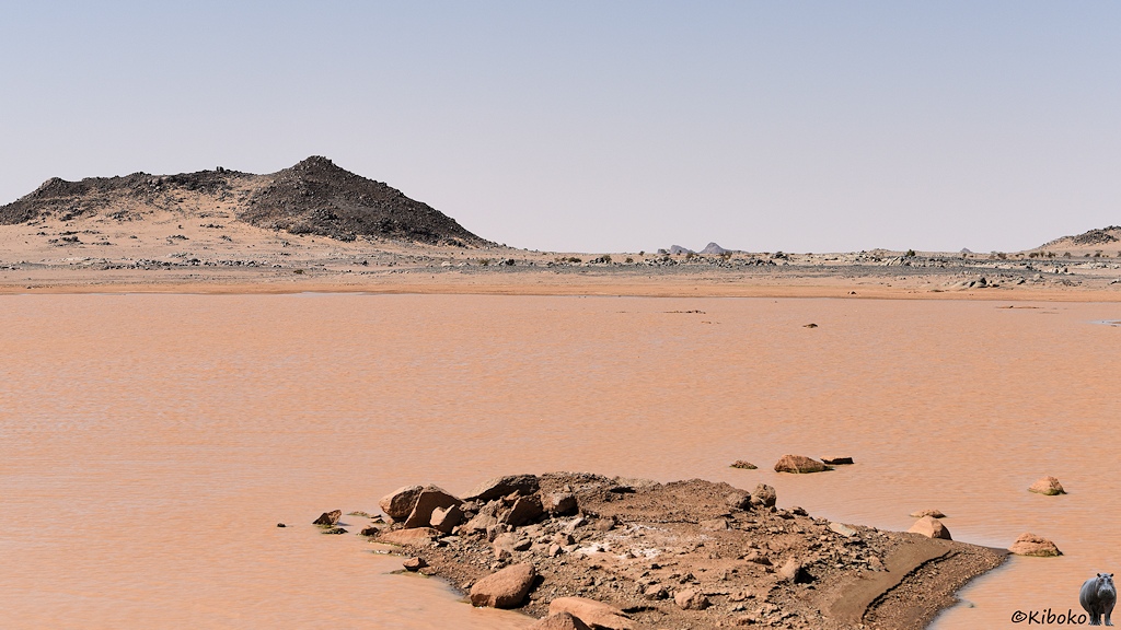 Das Bild zeigt einen See mit braunem Wasser. Im Vordergrund ragt eine Insel aus braunen Steinen und braunem Sand. Im Hintergrund sind ein Hügel mit schwarzen Steinen und eine Felsebene mit grauen Steinen.