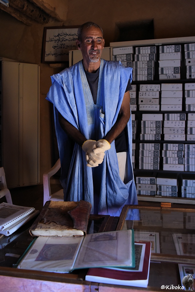 Das Bild zeigt Mann mit weißem Bart und lichtem Haar in einem blauen Gewandt und weißen Baumwollhandschuhen. Er steht vor einem offenen Stahlschrank mit beschrifteten weißen Kartons. Im Vordergrund liegt ein dickes, altes Buch.