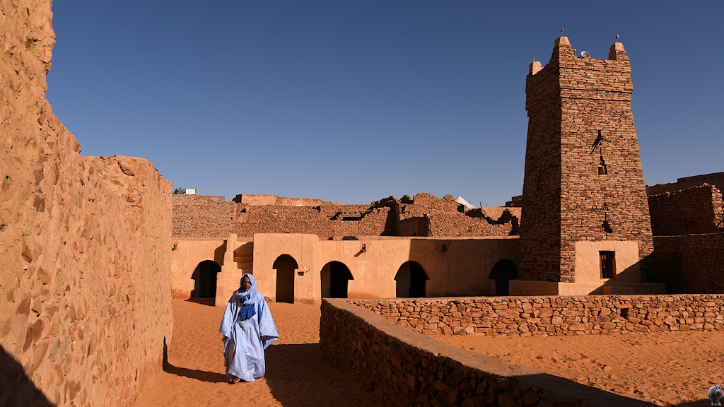 Das Bild zeigt den Innenhof einer Moschee mit einem sandigen Weg. Hinten ist wieder das verputzte Gebäude mit den pilzförmigen Eingängen. Am rechten Bildrand steht der viereckige Turm. Ein Mann im langen hellblauen Gewandt verlässt die Moschee.