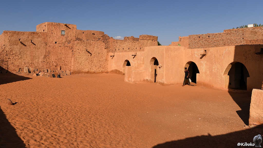Das Bild zeigt den sandigen Innenhof einer Moschee. Auf der rechten Seite ist ein einstöckiges Gebäude mit vier pilzförmigen Eingängen. Dahinter ist eine Wand aus rötlichen Natursteinen.