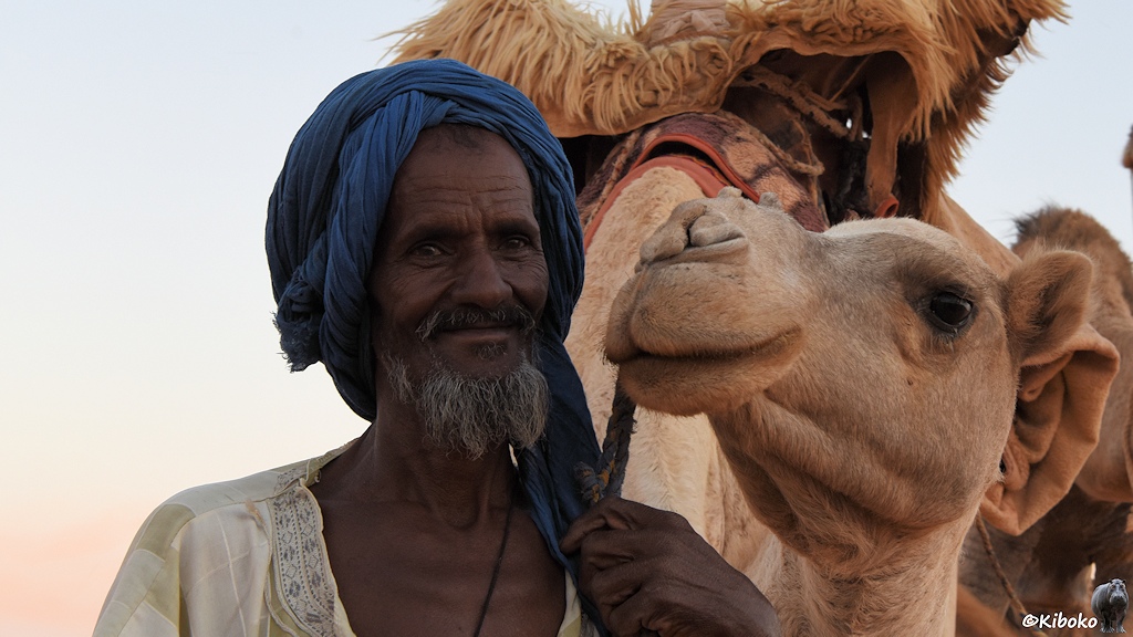 Das Bild zeigt einen älteren Mann mit weißem Bart und blauem Turban. Der Mann schaut mit großen Augen lächelnd in die Kamera. Rchts daneben ist der Kopf seines Kamels, das ebenfalls mit großen Augen und einem Lächeln in die Kamera schaut 