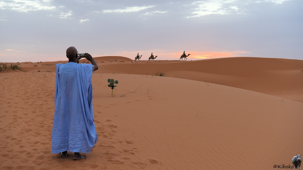 Das Bild zeigt einen Mann im hellblauem Gewandt der mit dem Handy fotografiert. Er steht in einer Wüste aus blass lachsfarbenen Sand. Am Horizont reiten drei Männer auf Dromedaren über einen Dünenkamm. Dahinter ist der Himmel einen orangen Streifen.