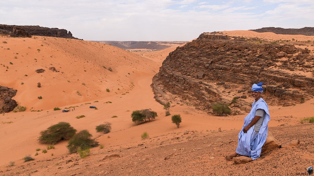 Das Bild zeigt Mann im hellblauem Gewand auf einem Felsen am einem Hang sitzen. Dahinter führt ein Tal mit lachsfarbenem Sand in das Bild. Das Tal ist rechts mit einer dunklen Felswand und links mit einem Hang aus Sand und einer abschließenden dunklen Felswand begrenzt. Im Hintergrund ist ein quergehendes breiteres Tal zu erahnen.